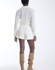 A model shown from the back wearing a white long-sleeve collared shirt and matching white shorts, paired with tan knee-high heeled boots, against a plain white studio background.