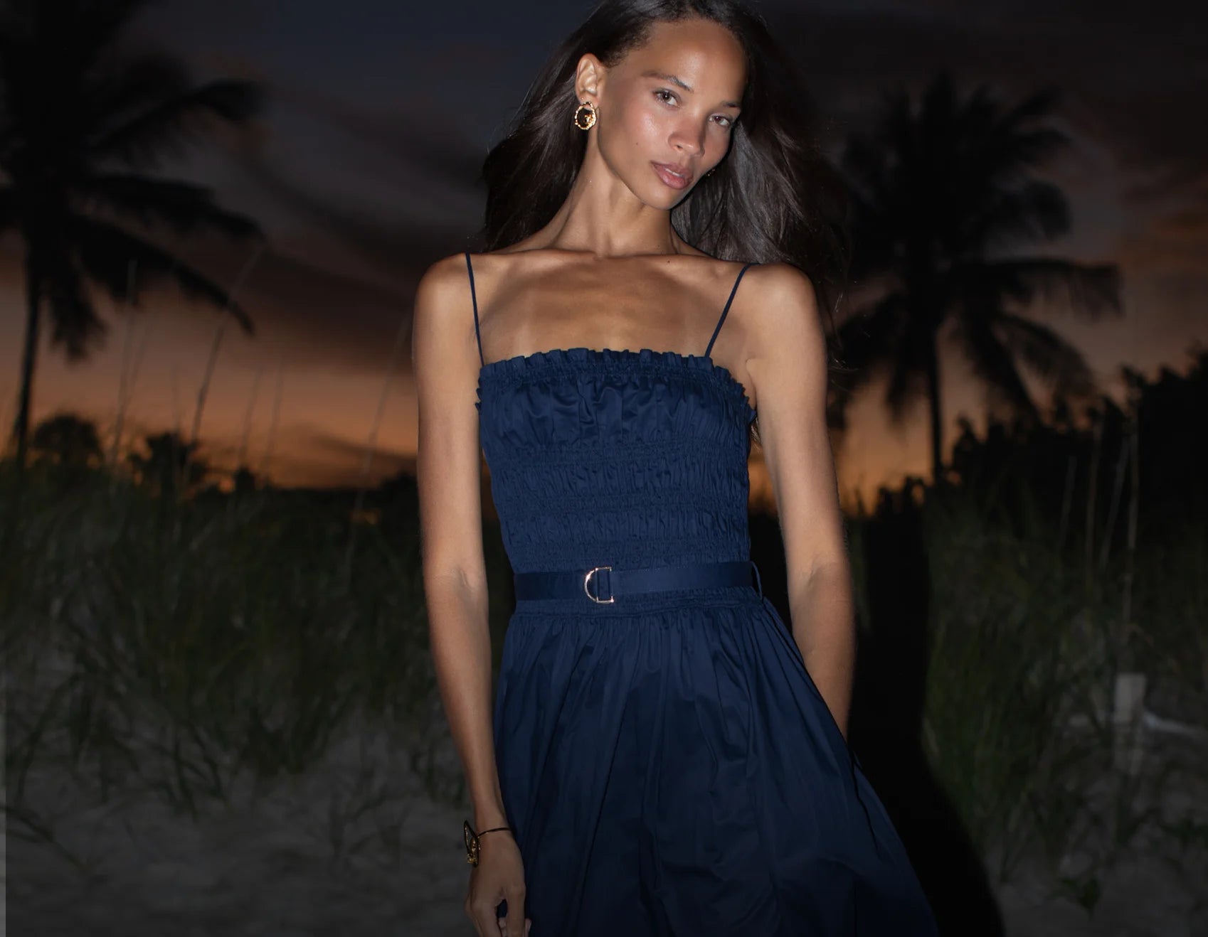 Woman in a blue dress standing against a dark, palm tree-lined background