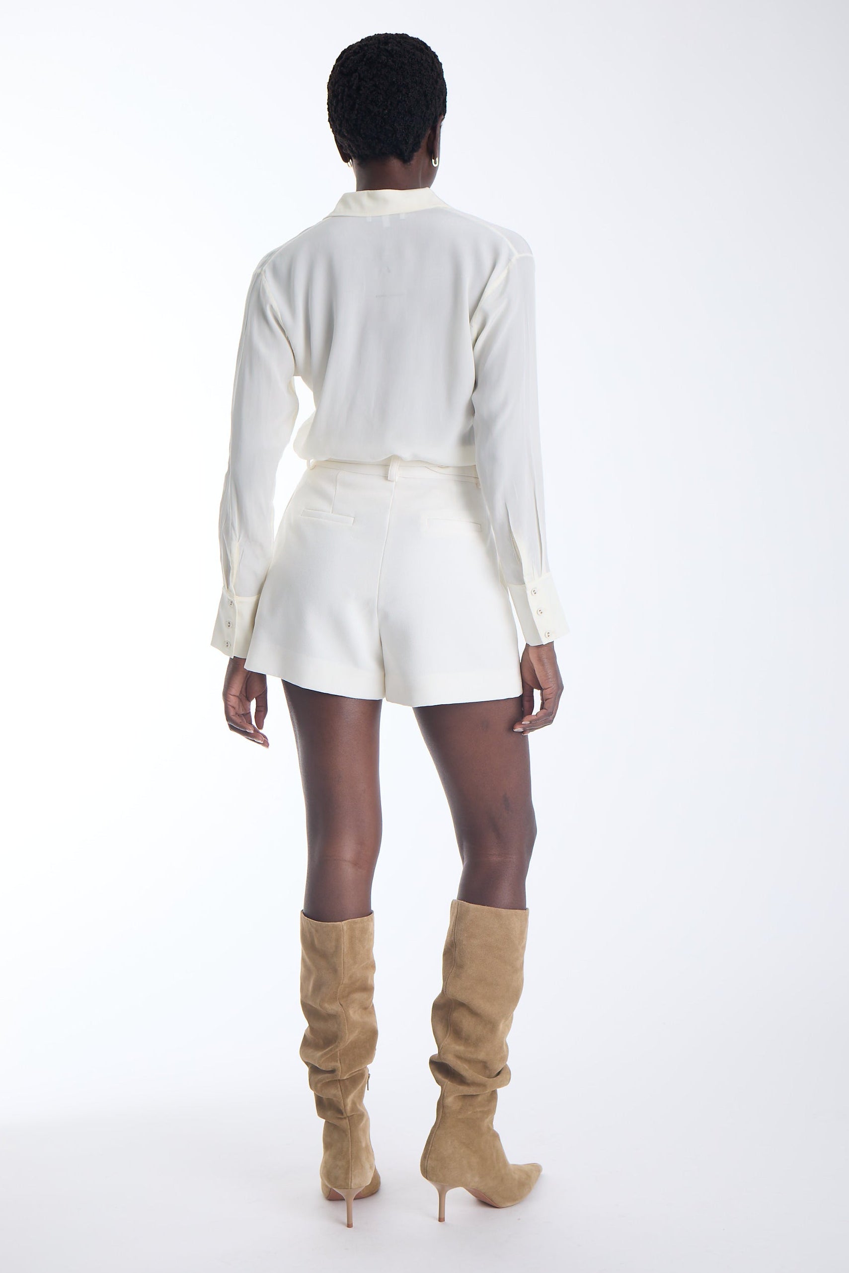 A model shown from the back wearing a white long-sleeve collared shirt and matching white shorts, paired with tan knee-high heeled boots, against a plain white studio background.