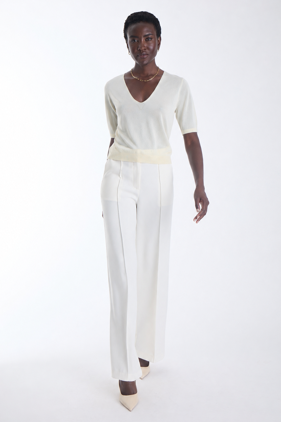 A model wearing a cream-colored short-sleeve knit top with a wide matching belt and high-waisted cream trousers, paired with beige pointed-toe heels, against a solid white studio background.